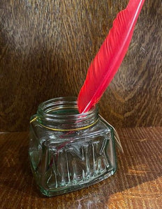 Image shows a glass pen storage pot that resembles an old empty jar of ink with the word ink stamped into the glass on one side. There is a few feather quill pen sitting in the jar.