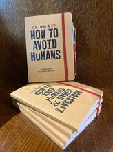 Load image into Gallery viewer, Image shows a small stack of kraft notebooks with one displayed stood on top. The stack of notebooks bear the slogan 'Grimm & Co. HOW TO KEEP HUMANS AND OTHER LIVESTOCK' and the one on top says 'Grimm & Co. HOW TO AVOID HUMANS an exercise in seclusion and obscurity'. The top notebook has a Word Wand (Fancy) held in the elastic pen loop, sold separately.