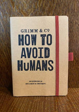 Load image into Gallery viewer, Image shows a kraft card notebook with red elastic closure displaying the slogan 'Grimm & Co. HOW TO AVOID HUMANS and exercise in seclusion and obscurity'. The notebook is A5 size and has a red elastic pen loop.