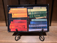 Load image into Gallery viewer, Image of an open Twists & Tales box on a display stand, showing the card categories within. There are four sets of cards decorated to look like books on a shelf, and they are colour coordinated. Blue cards for Objects, red cards for Characters, green cards for Settings, and yellow cards for Plot.