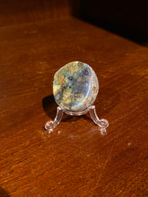 Load image into Gallery viewer, Image shows a labradorite palmstone with rough outer edge and gleaming colours inside, stood on a plastic stand.