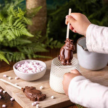 Load image into Gallery viewer, Image shows a hand holding the Gruffalo-shaped milk chocolate stirrer spoon over a glass of milk with a bowl of marshmallows on the wooden board next to them and green ferns in the background.