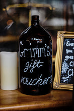 Load image into Gallery viewer, Image of a brown glass bottle with the words Grimm's Gift Vouchers written on in white chalk pen. Bottle is on a counter with a candle.