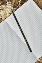 Load image into Gallery viewer, Image of a Word Wand, otherwise known as a black pencil with silver printed writing along the length and a black eraser at the top. The lettering on this side of the pencil reads 'Mistakes' with an arrow pointing towards the eraser, and 'Magic' with an arrow pointing towards the lead. The Word Wand rests on a lined kraft note book.