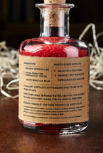 Load image into Gallery viewer, Image shows a bottle of Middle Aged Vitriol, otherwise known as red, scented bath salts in a glass bottle with cork lid and a kraft paper label. Detail of back of label showing faux ingredients and side effects for mortals.