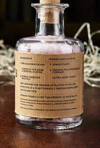 Image shows the back of a bottle of Grandma's Scabs potion, a blass bottle with cork containing pale pink bath salts and dried rose petals. Bottle is wrapped with a kraft paper label, showing the faux ingredients and side effects.