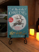 Load image into Gallery viewer, Image of the paperback book called A Boy Called Christmas stood on a book stand with candles