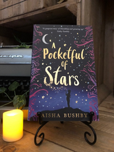 Load image into Gallery viewer, Image of the front cover of the paperback book A Pocketful of Stars stood on a book stand with candles