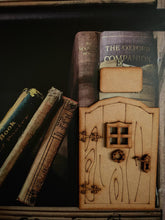 Load image into Gallery viewer, Image shows a square edition fairy door stuck against some books on a shelf. The door is unpainted, and has the extras of a window frame, door handle, small floral decoration, and small sign all displayed.
