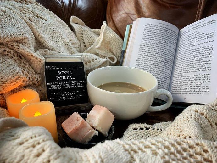 Image showing a white blanket on a brown sofa with an open book and a tray holding two battery operated candles, a cup of coffee and a mini bowl containing two wax melts and the Cosy Book Nook Scent Portal sat next to them.