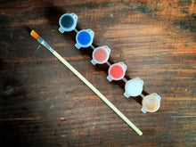 Load image into Gallery viewer, Close up image of the paintbrush and paints included in the fairy door pack. The colours are black, brown, orange, blue, gold, and white.