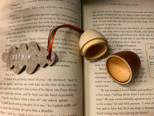 Load image into Gallery viewer, Image of A Kiss, a wooden acorn-shaped hanging ornament with a natural wood finish on the top half, and rose gold painted bottom half. Acorn splits in two with a hollow inside to hide messages or trinkets. Attached to the hanging loop is a grey felt oak leaf with the words A Kiss printed in white. Decoration is shown displayed on the pages of Peter Pan.