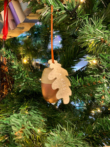 Image shows A Kiss wooden acorn-shaped hanging decoration displayed hung from a Christmas tree branch. A grey felt oak leaf is attached to the hanging loop. printed with the words A Kiss.
