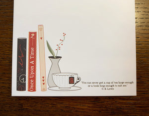 A close up image of the illustration and quote printed at the bottom of the notepad. There are three books stacked next to one another, showing the spines. One is navy blue with a 'squiggle' title, the next is red with once upon a time' along the spine, and the third is pale pink/beige with red hearts on the spine.  Next to the books is a grey vase, with a leaf and berry-like flowers inside, and a white teacup with a teabag tag hanging on the outside. The tag has the Grimm & Co. logo on it.