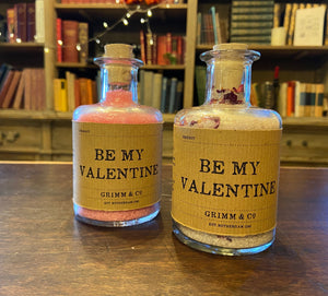 Image of two glass potion bottles filled with pink bath salts, one including rose petals. Both are labeled: Be My Valentine.
