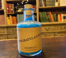 Load image into Gallery viewer, Image of a glass potion bottle filled with deep blue bath salts. The bottle label reads: Congratulations!