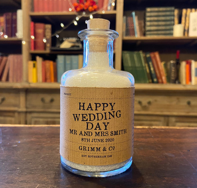 Image of a glass potion bottle filled with white bath salts. The bottle label reads: Happy Wedding Day Mr and Mrs Smith, 8th June 2020.