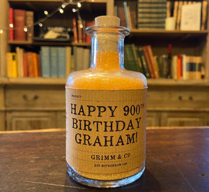 Image of a glass potion bottle filled with orange bath salts. The bottle label reads: Happy 900th Birthday Graham!