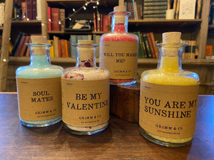 Image of four personalised potion bottles, otherwise known as scented bath salts in a glass bottle with cork and a brown kraft label. From left to right - Blue Potion label reads: Soul Mates. Pink and rose petal  Potion label reads: Be My Valentine. Red Potion label reads: Will You Marry Me? Yellow Potion label reads: You Are My Sunshine.