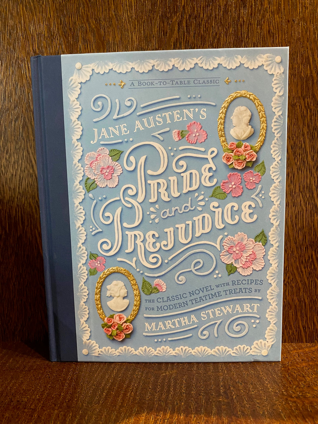 Image shows the front cover of the book Pride & Prejudice - A Book to Table Classic. The cover is pastel blue with a navy blue linen spine. There are illustrations of cameos of MR. Darcey and Elizabeth Bennett and icing sugar flowers decorating the cover. The text is white.