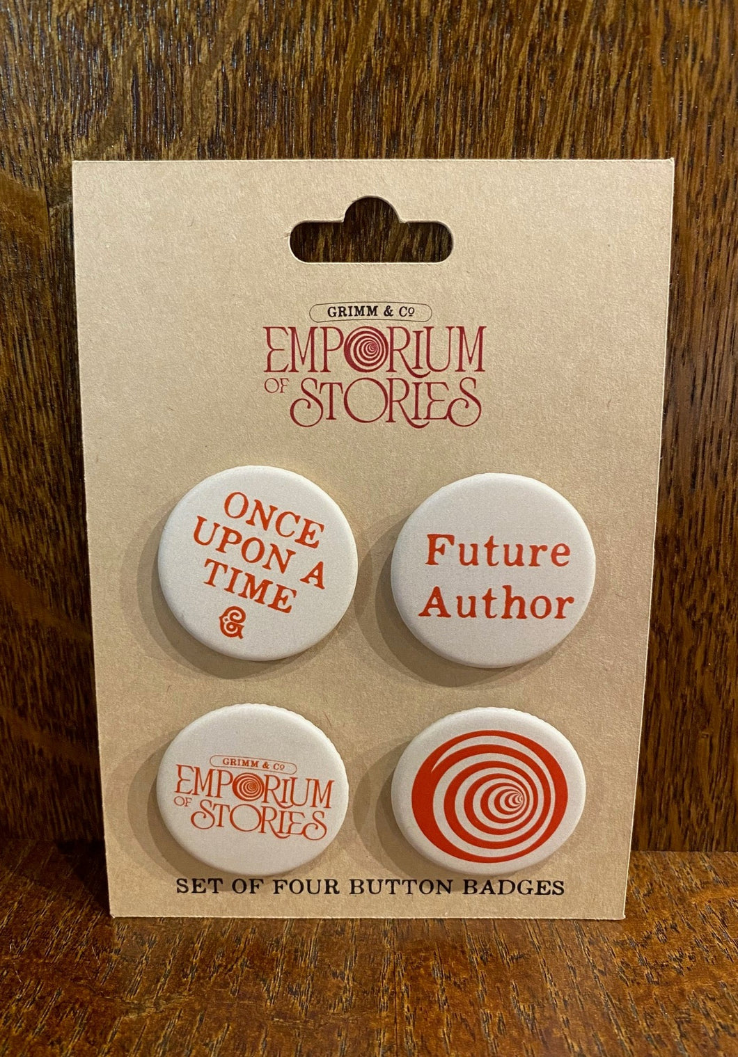 Image shows a set of four button badges set on a kraft card. Designs feature a cream background and red text. Text reads, 'Future Author', 'Once Upon A Time', the Grimm & Co. Emporium of Stories logo and a red spiral swirl from the Emporium logo.