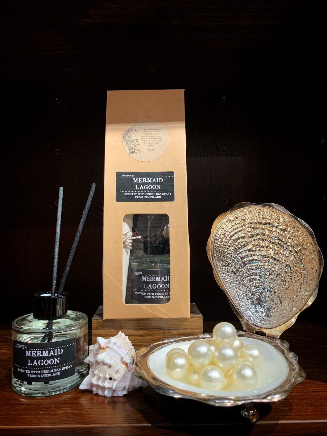 Image shows an open reed diffuser called Mermaid Lagoon scented with Sea Spray and displayed with a metal oyster shell and pearls and a white shell. 