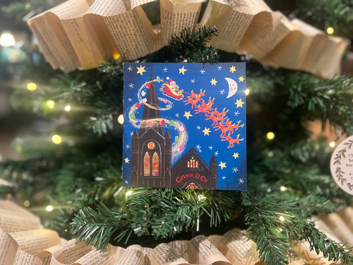 Image shows a Christmas card sat in a Christmas tree. Design features Santa flying over Grimm & Co's Emporium of Stories. Card designed by Lydia Monks.