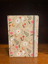 Load image into Gallery viewer, Image shows an A5 hardback notebook with a repeating design on a beige background featuring mustard crescent moons, daisies, red toadstools, pink tulips and green fairy wings.