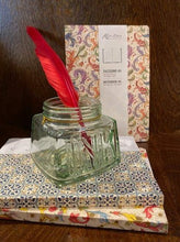 Load image into Gallery viewer, Image shows a small stack of two notebooks with a third stood behind, and a glass ink jar sat on top of the stack with a red feather quill inside. The notebooks are all various patterns and colours in decorative Italian paper with gold powder finish.