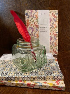 Image shows a small stack of two notebooks with a third stood behind, and a glass ink jar sat on top of the stack with a red feather quill inside. The notebooks are all various patterns and colours in decorative Italian paper with gold powder finish.