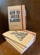 Load image into Gallery viewer, Image shows a small stack of kraft notebooks with one displayed stood on top. The stack of notebooks bear the slogan &#39;Grimm &amp; Co. HOW TO KEEP HUMANS AND OTHER LIVESTOCK&#39; and the one on top says &#39;Grimm &amp; Co. HOW TO AVOID HUMANS an exercise in seclusion and obscurity&#39;. The top notebook has a Word Wand (Fancy) held in the elastic pen loop, sold separately.