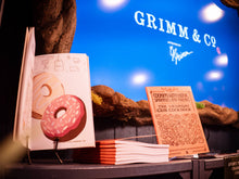 Load image into Gallery viewer, Image shows the display of Cooking With Magic, Sprinkles And Friends cookbook written by the Saturday Club children. One book stands open showing an illustration of donuts with coloured icing and sprinkles.