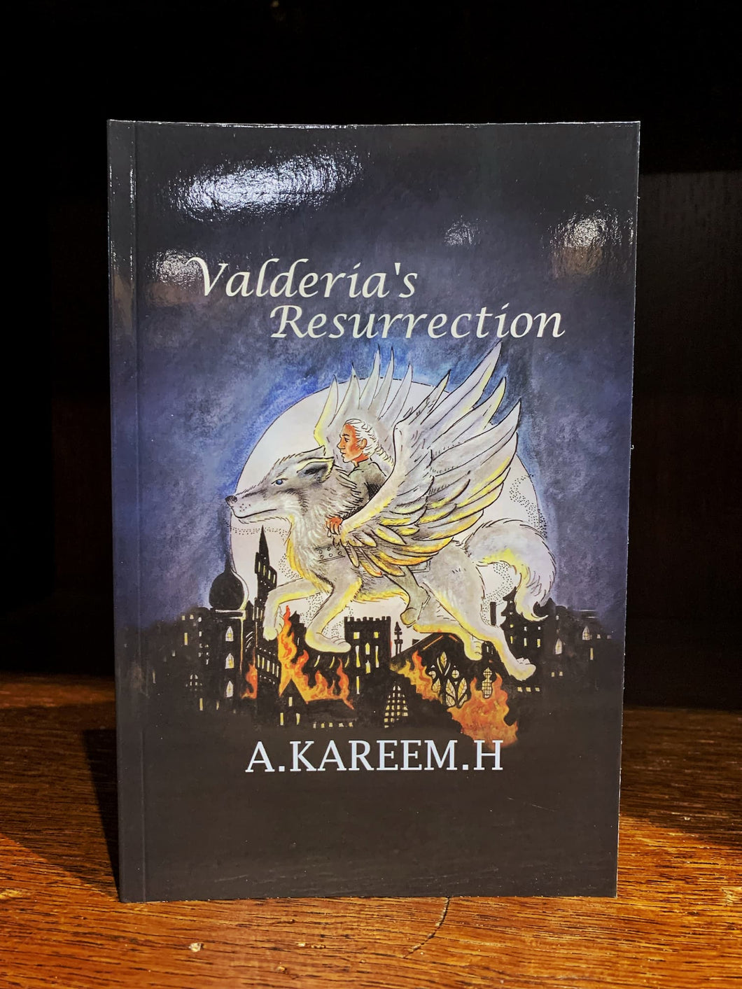 Image shows the cover of the ovella Valderia's Resurrection. The cover has an illustration of a man with white hair riding a winged wolf, and they are flying over the silhouette of a burning city, in front of a full moon. 