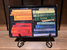 Load image into Gallery viewer, Image of an open Twists &amp; Tales box on a display stand, showing the card categories within. There are four sets of cards decorated to look like books on a shelf, and they are colour coordinated. Blue cards for Objects, red cards for Characters, green cards for Settings, and yellow cards for Plot.