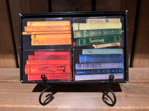 Image of an open Twists & Tales box on a display stand, showing the card categories within. There are four sets of cards decorated to look like books on a shelf, and they are colour coordinated. Blue cards for Objects, red cards for Characters, green cards for Settings, and yellow cards for Plot.
