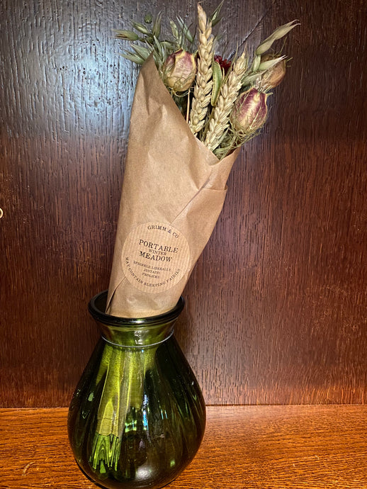 Image shows a bouquet of dried flowers in shades of red, green and natural wrapped in kraft paper in a green glass vase.