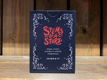 Load image into Gallery viewer, Image showing the front of the cardboard box for the card game Stems of a Story, created by children and young people during workshops at Grimm and Co. It is a small, dark blue box, with white leafy illustrations in each corner, and red and white text.