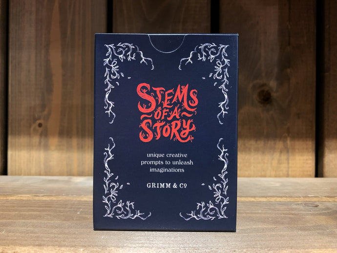Image showing the front of the cardboard box for the card game Stems of a Story, created by children and young people during workshops at Grimm and Co. It is a small, dark blue box, with white leafy illustrations in each corner, and red and white text.