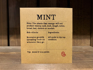 Image shows a packet of Mint seeds. It is a square, flat, kraft brown paper packet., with black text printed in Appareo font. It says Mint at the top, with magical ingredients and side effects printed underneath, and a magical 'caution' at the bottom.