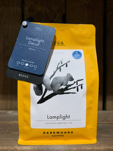 Image of the Roasted Magic Beans (Whole) - Lamplight Decaf coffee beans. The image shows a bright yellow packet with a black and white drawing of a squirrel on a branch. There are two labels attached to the top left corner of the packet with the name of the coffee, the fact that it is decaf, and the label underneath states that they are beans.