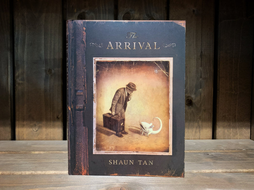 Image of the front cover of The Arrival. It is designed to look like a distressed, old leather book. There is an illustration of a sepia photograph that shows a man in a suit holding a carry-case peering over to look at a strange white creature.