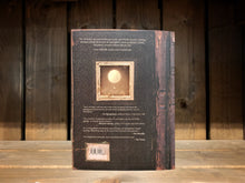 Load image into Gallery viewer, Image shows the back cover of The Arrival. the distressed old leather book theme continues, and there is an illustration of a small square sepia photograph. the photograph shows a glowing balloon with a small container hanging underneath. 