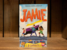 Load image into Gallery viewer, Image of the front cover of the book Jamie. The illustration on the front shows three children/young people stood on the roof of Nottingham town hall holding a non-binary flag, while a crowd with various LGBT+ pride flags is below. The title is written across the top in a large, cartoonish-style font in bright orange. 