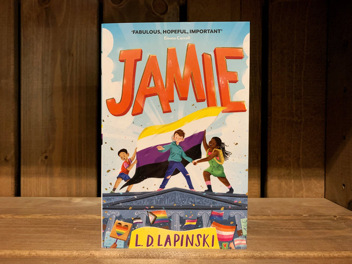 Image of the front cover of the book Jamie. The illustration on the front shows three children/young people stood on the roof of Nottingham town hall holding a non-binary flag, while a crowd with various LGBT+ pride flags is below. The title is written across the top in a large, cartoonish-style font in bright orange. 