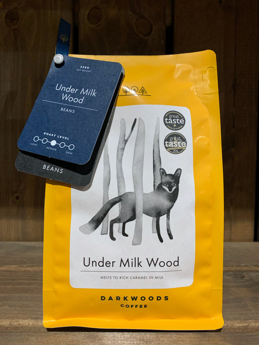 Image of the Roasted Magic Beans (Whole) - Under Milk Wood coffee beans. The image shows a bright yellow packet with a black and white drawing of a fox walking through trees. There are two labels attached to the top left corner of the packet with the name of the coffee, the fact that it is decaf, and the label underneath states that they are beans.