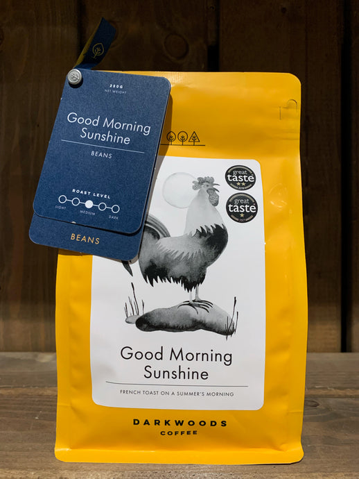 Image of the Roasted Magic Beans (Whole) - Good Morning Sunshine coffee beans. The image shows a bright yellow packet with a black and white drawing of a rooster. There are two labels attached to the top left corner of the packet with the name of the coffee, the fact that it is decaf, and the label underneath states that they are beans.