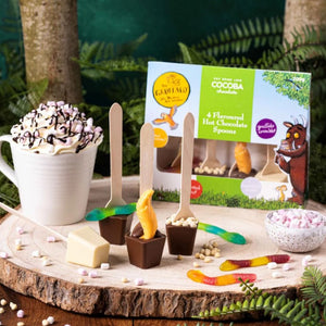 Image shows the 4 pack of flavoured hot chocolate stirrer spoons each with a difference character theme. Boxed stirrers sit in the background and each spoon sits in the foreground on a wooden log board with a mug of hot chocolate and cream sat next to them.