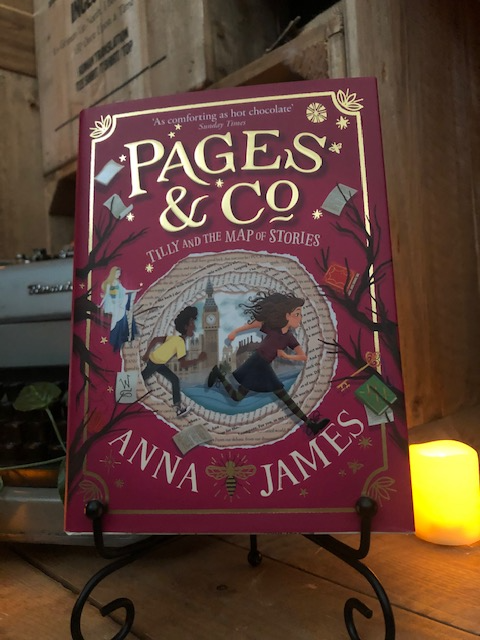Image of the front of the hardback book Pages and Co: Tilly and the Map of Stories written by Anna James and illustrated by Paola Escobar. Displayed on a book stand with candles.