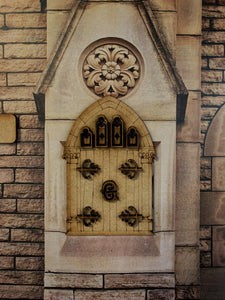 Image shows a special Grimm edition fairy door stuck onto a decorative stone background. All door accessories are also cut out and stuck on - window frames, decorative hinges, and the center Grimm insignia.