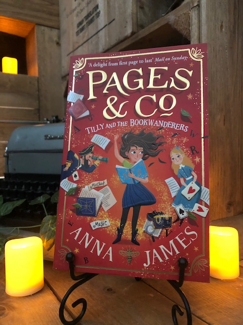 Image of the front of the paperback book Pages & Co: Tilly and the Bookwanderers written by Anna James and illustrated by Paola Escobar. Displayed on a book stand with candles.
